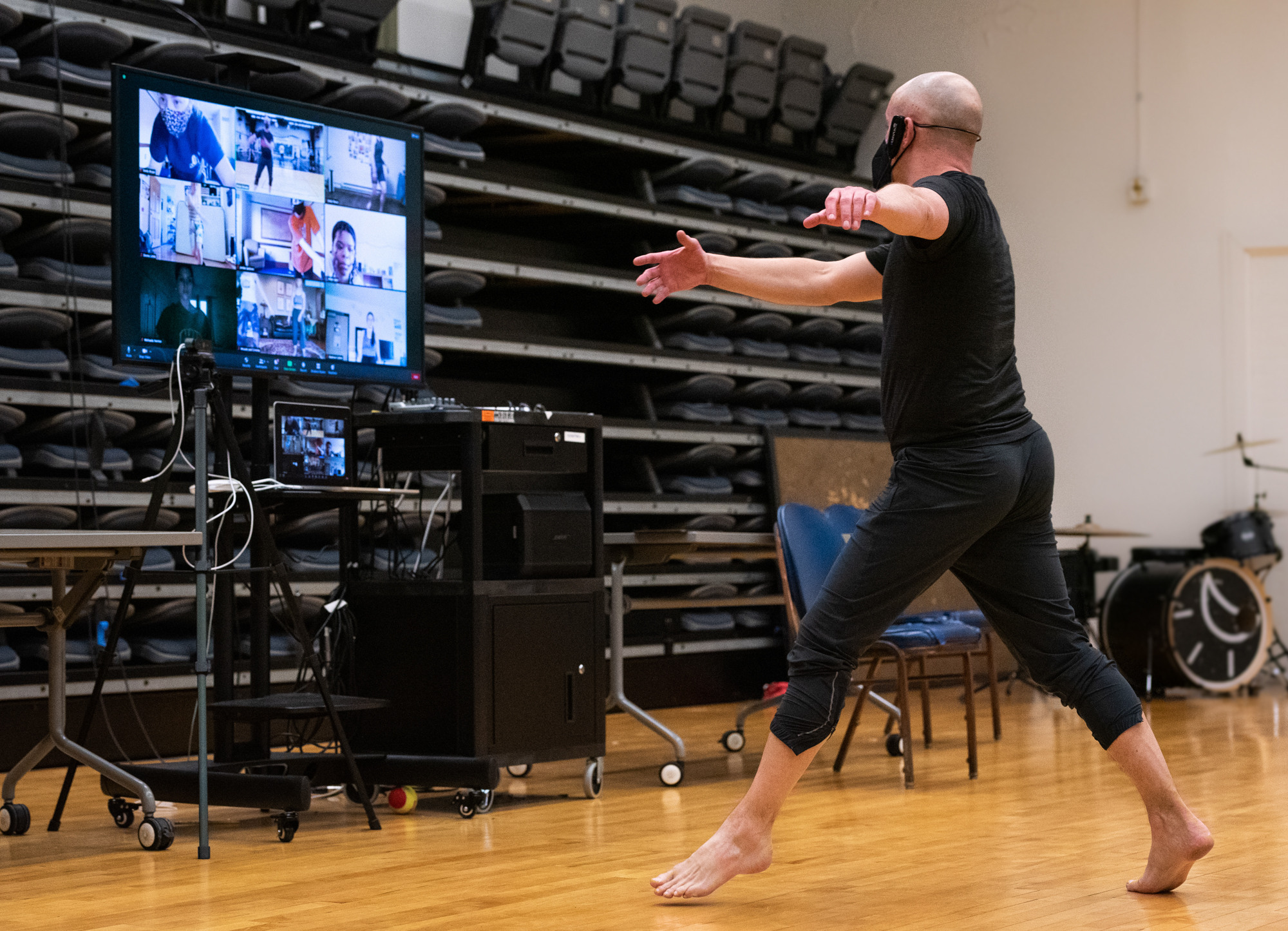 A man dances barefoot in front of a large screen that shows video call participants as part of remote dance practice