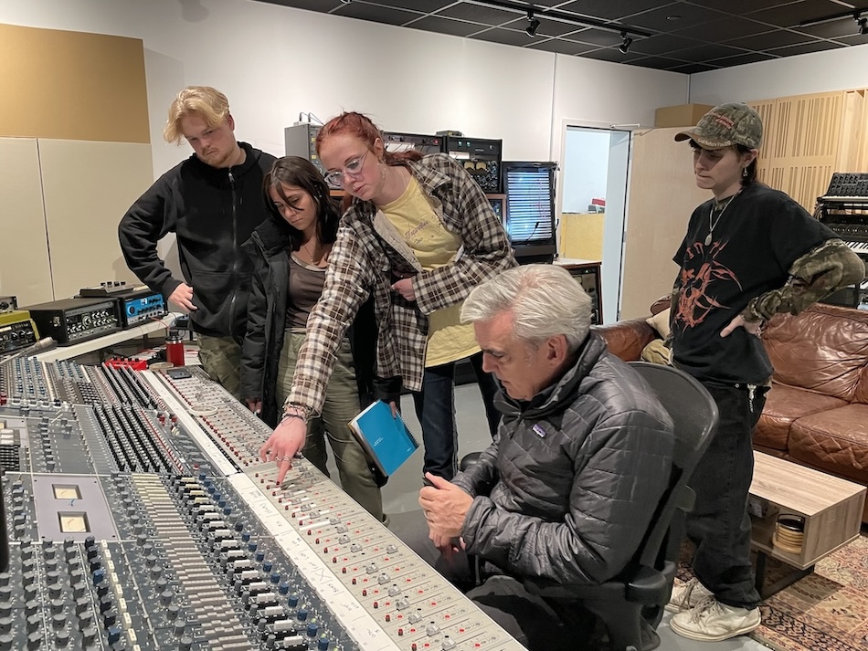 Students stand behind a producer at a sound board; one reaches over to point something out