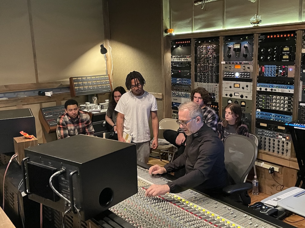 Students stand behind a sound engineer as he works at the large soundboard in front of him