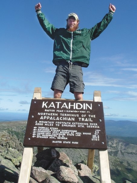 Adam Jones posing triumphantly over a sign reading Mount Katahdin, Maine.