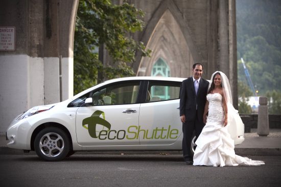 A man in a suite and a woman in a wedding dress stand next to a white vehicle bearing the words 