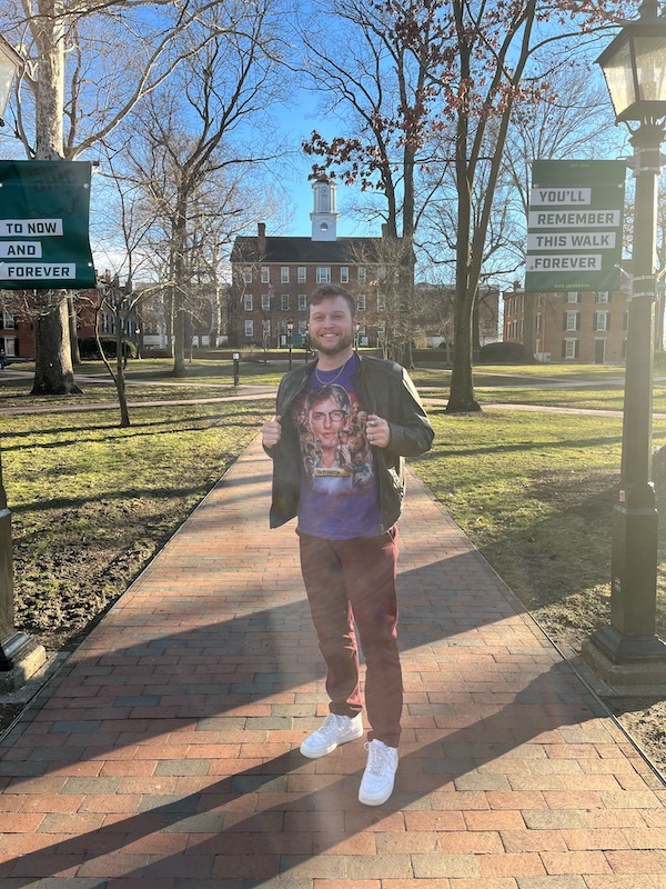 A man smiles at the camera from a brick path on OHIO's College Green, with Cutler Hall behind him