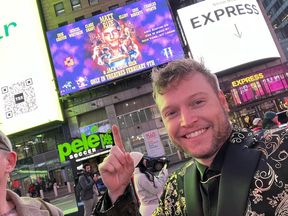 A man takes a selfie pointing to a large movie screen in Times Square