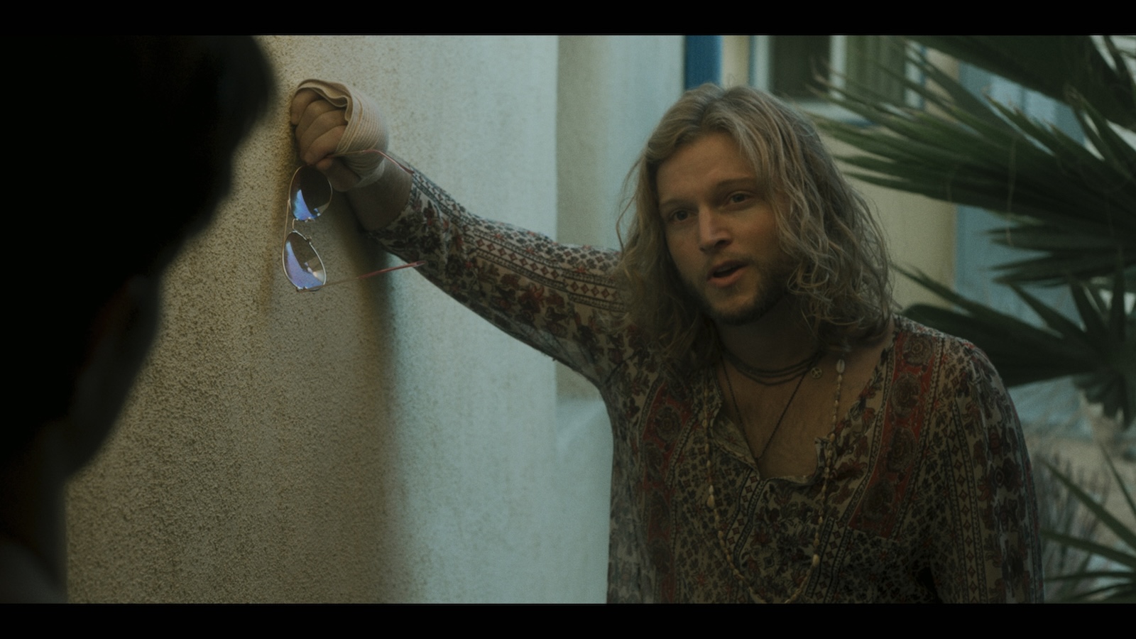 A man with shoulder-length hair leans against a wall, speaking to someone who's back is to the camera