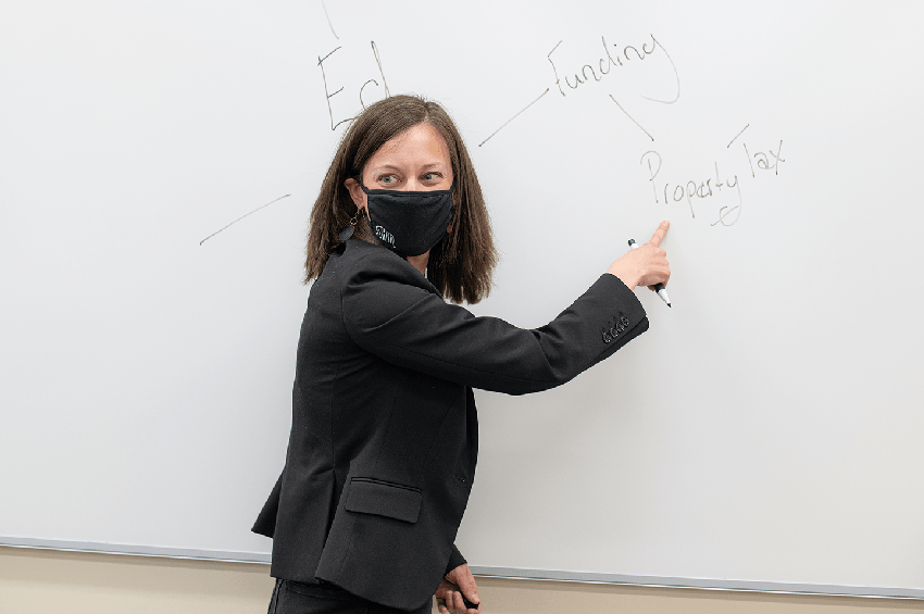 Women in a black suit jacket and black COVID-19 mask pointing to a white board while holding a erasable marker