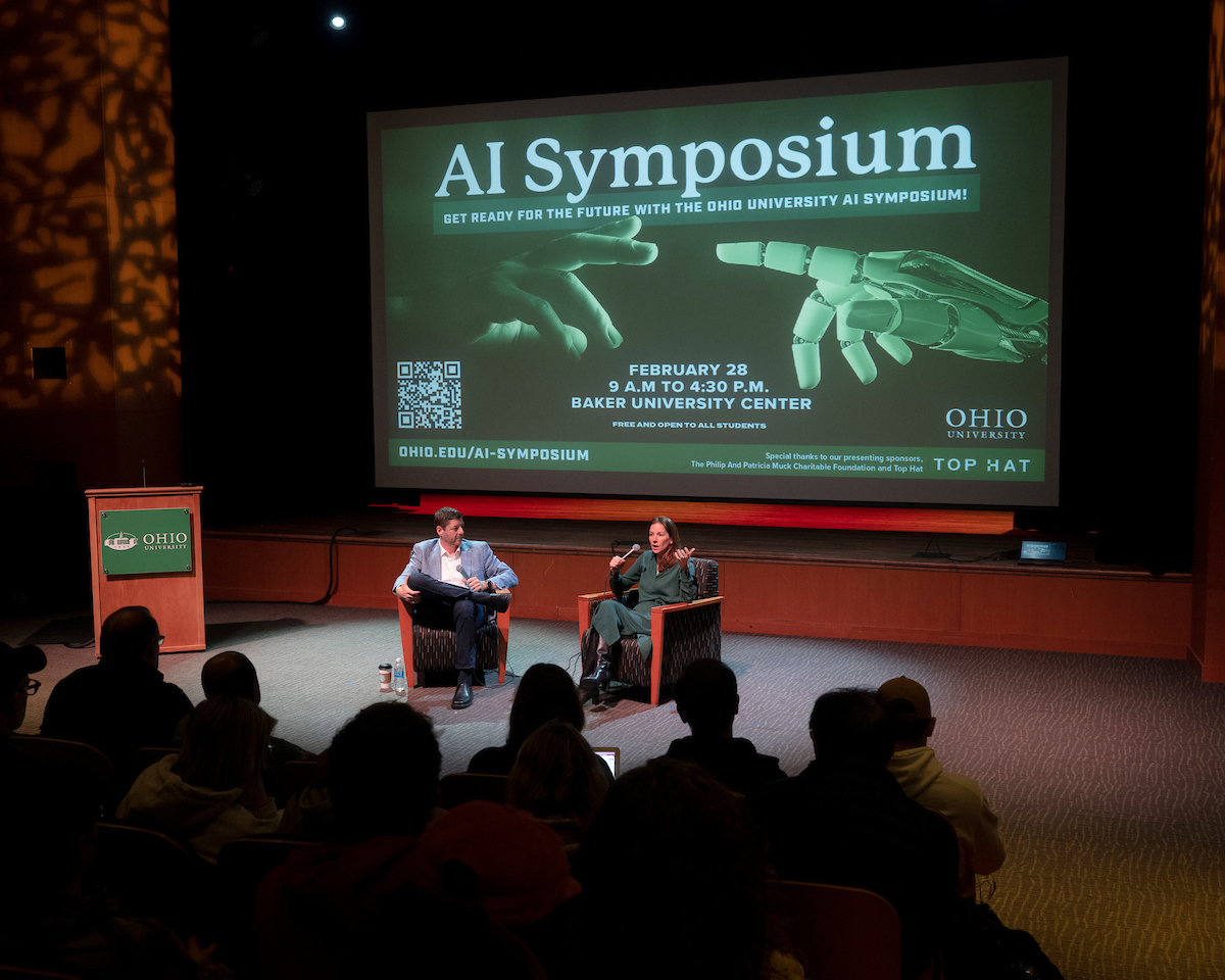 Two speakers sit in front of an audience in front of a large screen reading "AI Symposium" 