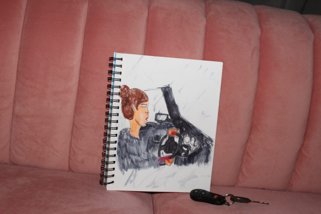 Upon a pink, plush couch lies two items: one is a set of car keys and the other is a sketchbook opened to a page with a drawing. The drawing, created with pen and markers, features a brunette person driving in a vehicle with a grey interior. This person has blood coming from their nose and on their hands. The person is crying.