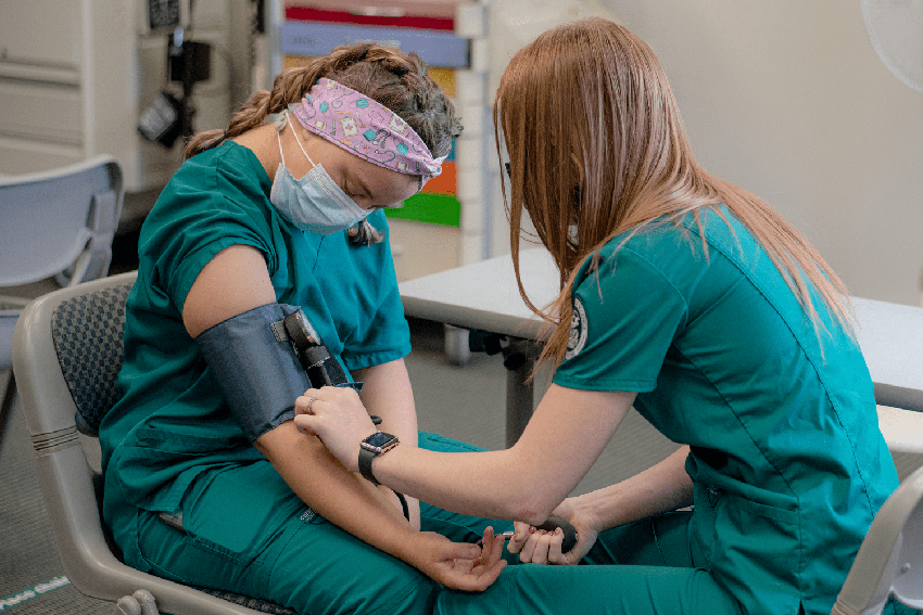 1 nursing student in green scrubs is checking the pulse of another nursing student in green scrubs. they are sitting down facing each other