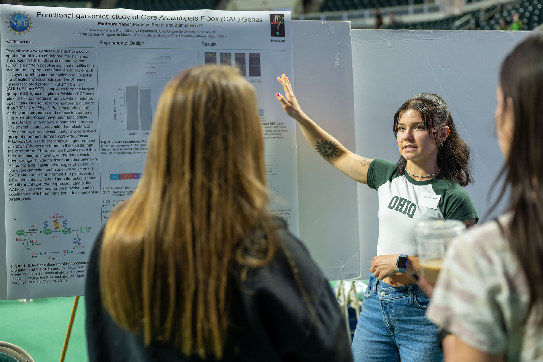a student points to her research presentation display while two observers look on