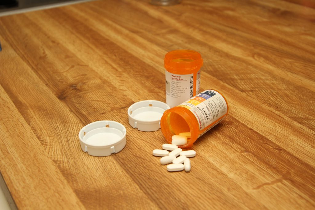 Two bottles of pills with their prescription labels sit on a wooden floor. One bottle is on its side, with eleven white pills spilling from it. The other bottle is upright. Both bottles are open and the caps for the bottles lay beside them.