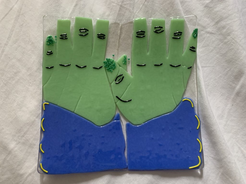 Glass art of two green hands with overlapping thumbs. The tips of the fingers are not visible, except for the thumbs and pinky fingers, which have dark green puffy dot paint covering the nails. The sleeves are blue with yellow accents. There is clear glass supporting the hands and arms.