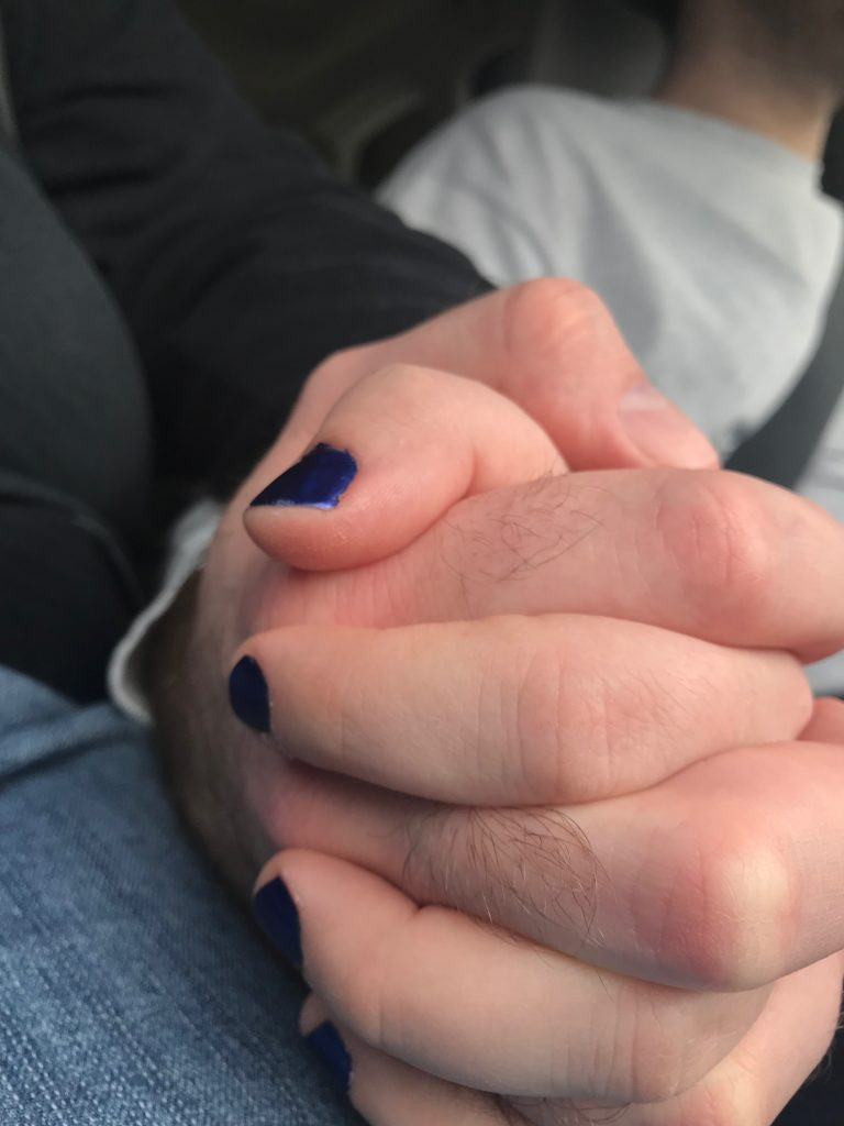 Two people hold hands, their fingers intertwined. One person wears blue fingernail polish. The hands are sitting on one person’s lap, and one appears to be driving as there is an out of focus portion of their shoulder and a seat belt in frame.