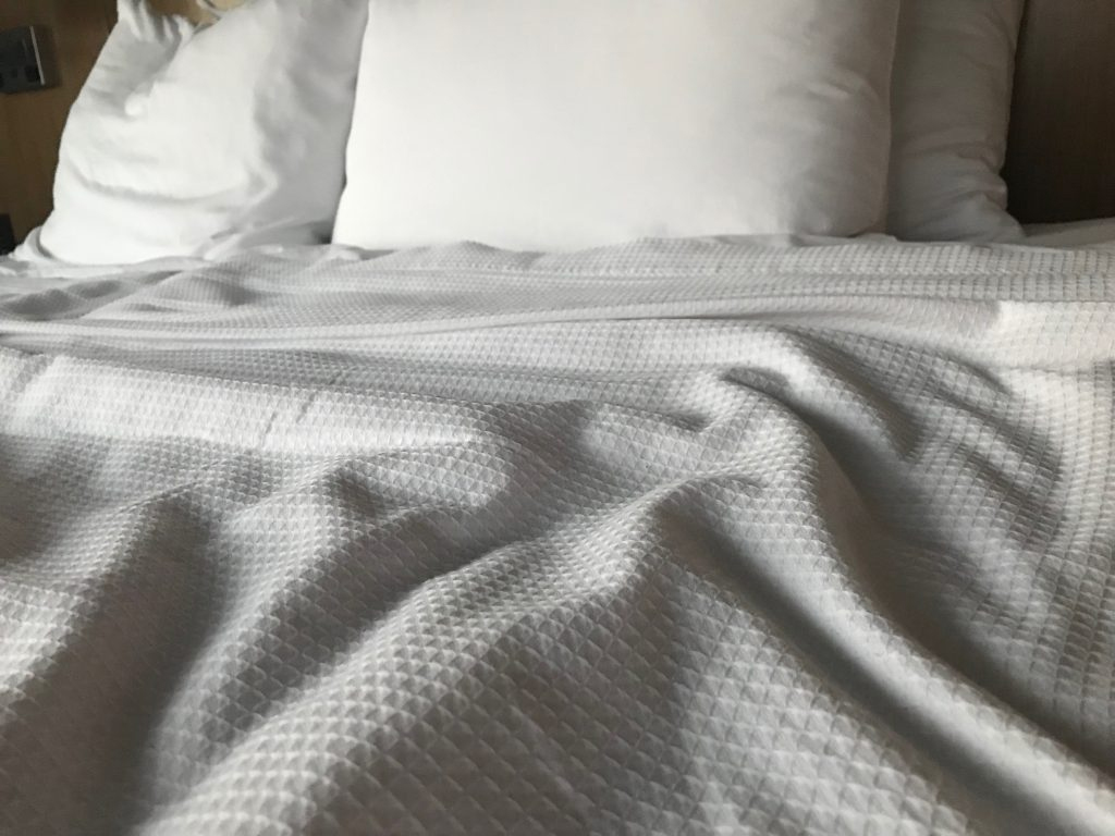 A bed with three pillows, but only one looks as though it has a head print from when it was used. The white sheets are rumpled, but pulled up to the pillows.