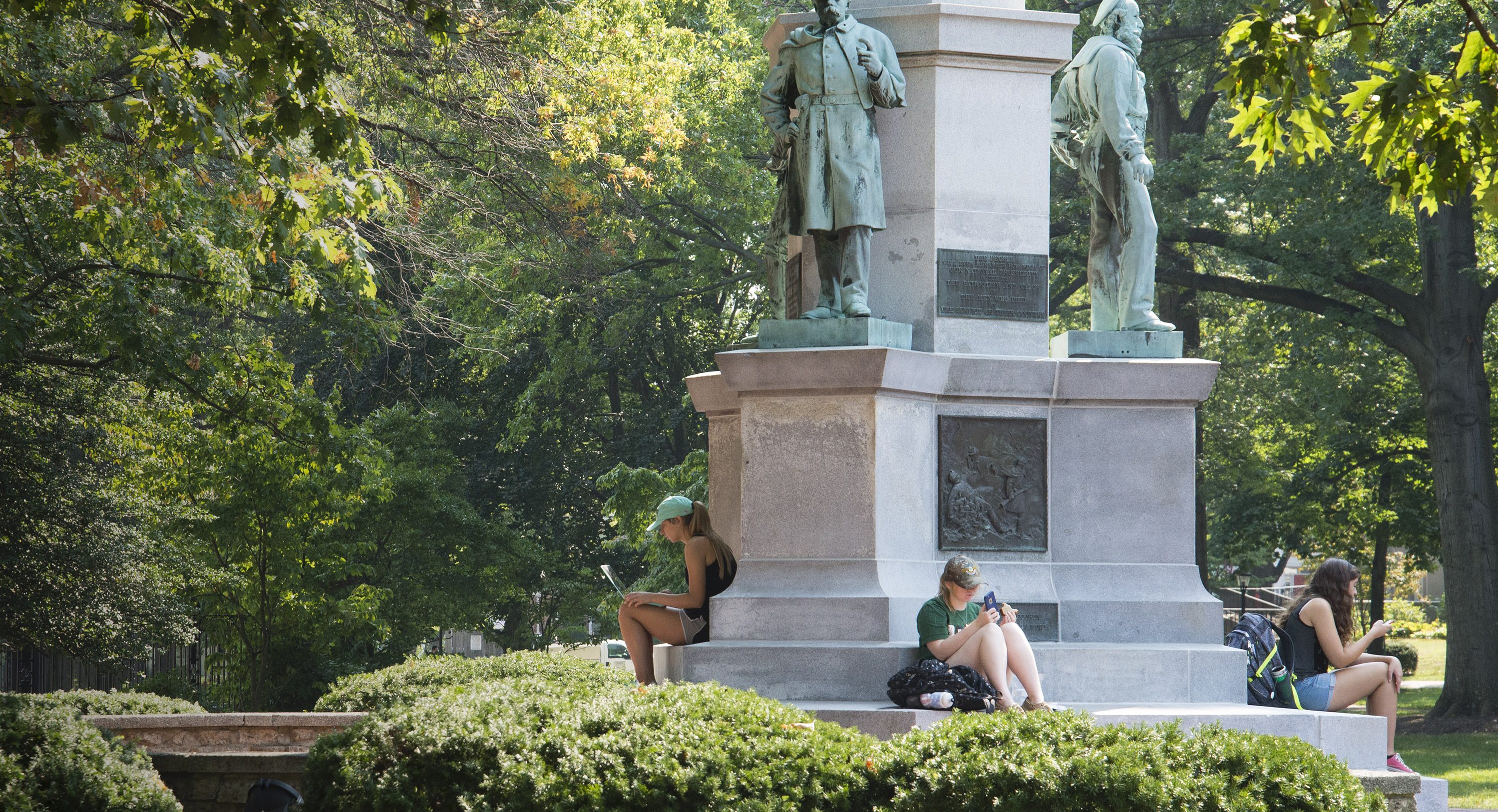 Three women students sit separately around the base of the Soldiers Monument on Ohio University Athens campus