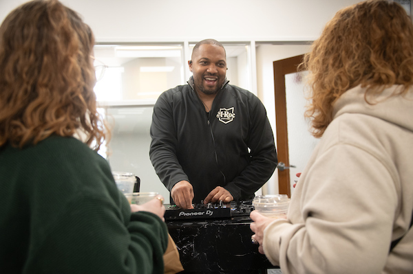 Smiling, a man speaks to two people with their backs to the camera