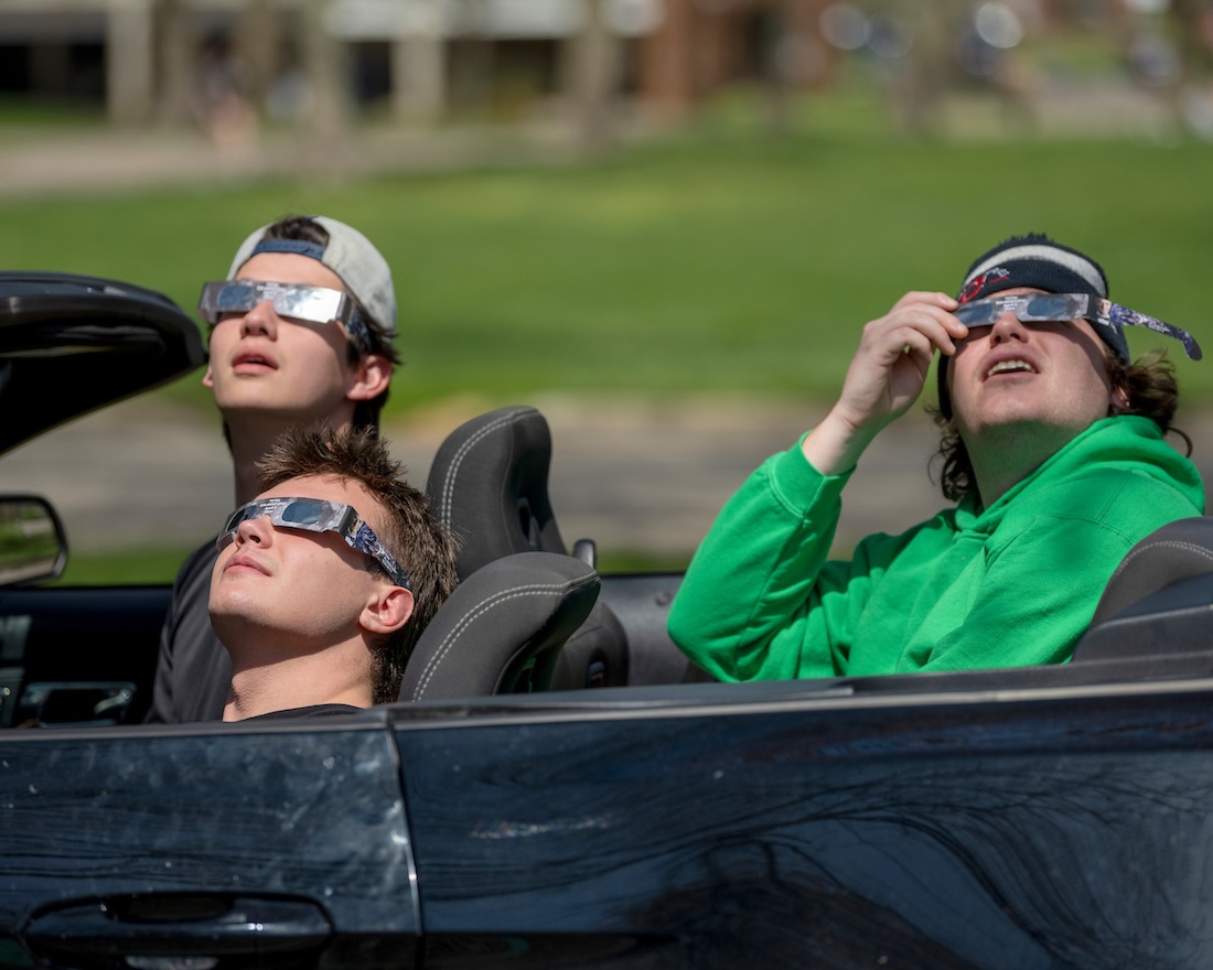 Three students wearing solar eclipse glasses look at the sky from a convertible