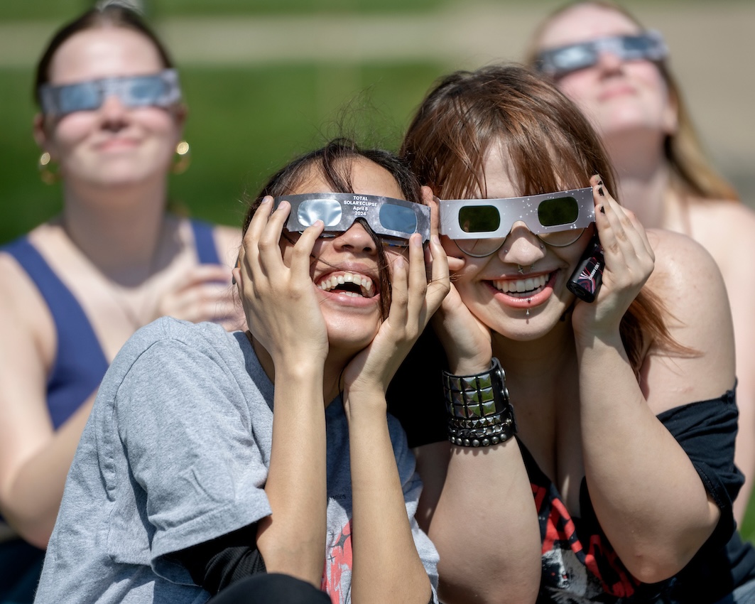 Two students wearing dark eclipse glasses sit close together, smiling broadly for the camera