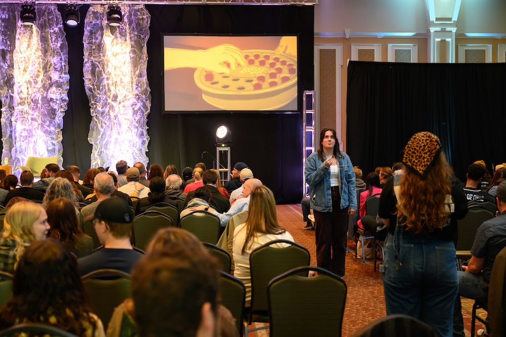 A crowd is pictured from the back of a crowded auditorium and a student speaks into a mini-microphone in an aisle while being filmed