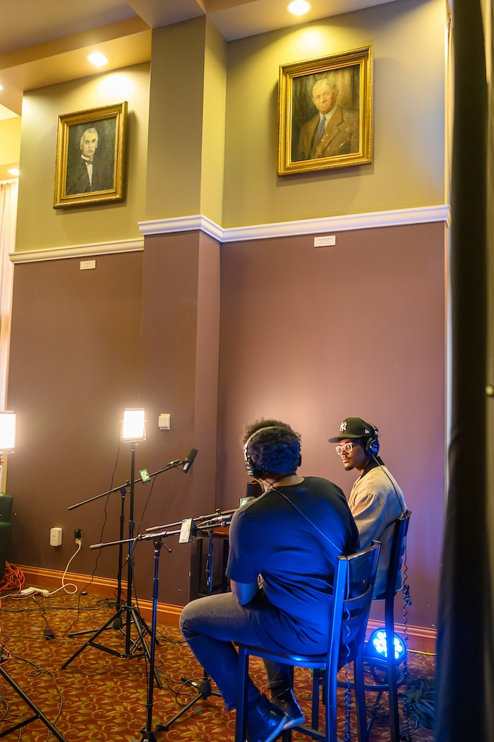 Pictured from behind, two people sit in front of microphones