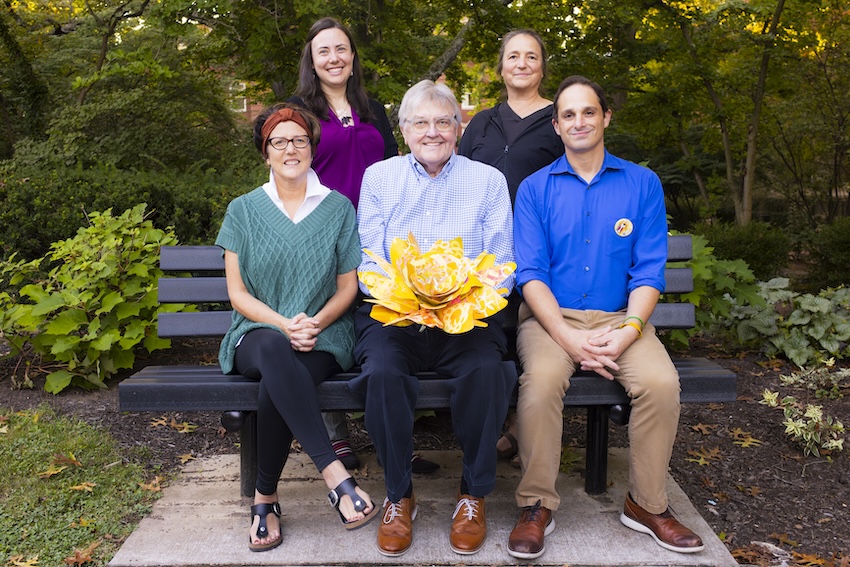 A group of five people is pictured, with the man in the middle holding a large yellow metal flower