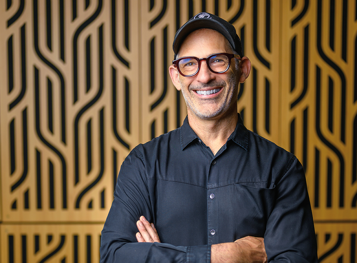 a man wearing a button-down shirt, baseball cap and glasses stands in front of a textured wall, smiling