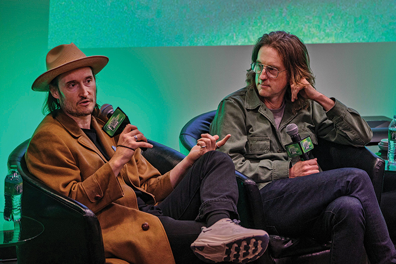 two members of the band My Morning Jacket sit in comfortable chairs on a stage, holding microphones in casual conversation
