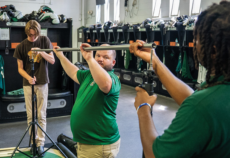 an Ohio University employee holds a crossbar while two students set it on tripods in a locker room 