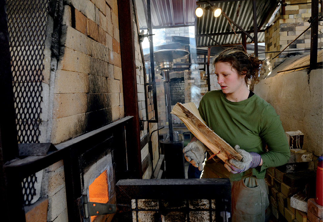 an undergraduate student wearing long sleeves and protective gloves prepares to load a log into a wood-burning kiln