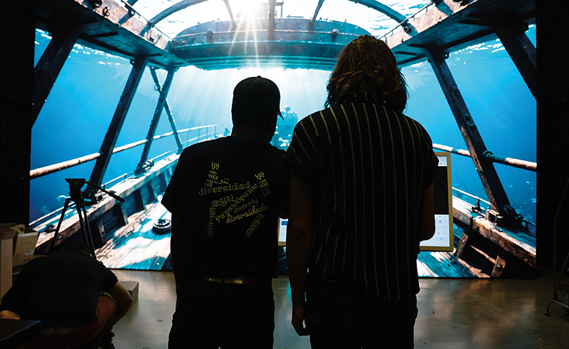 two people stand silhouetted against a screen displaying the view from an underwater submarine