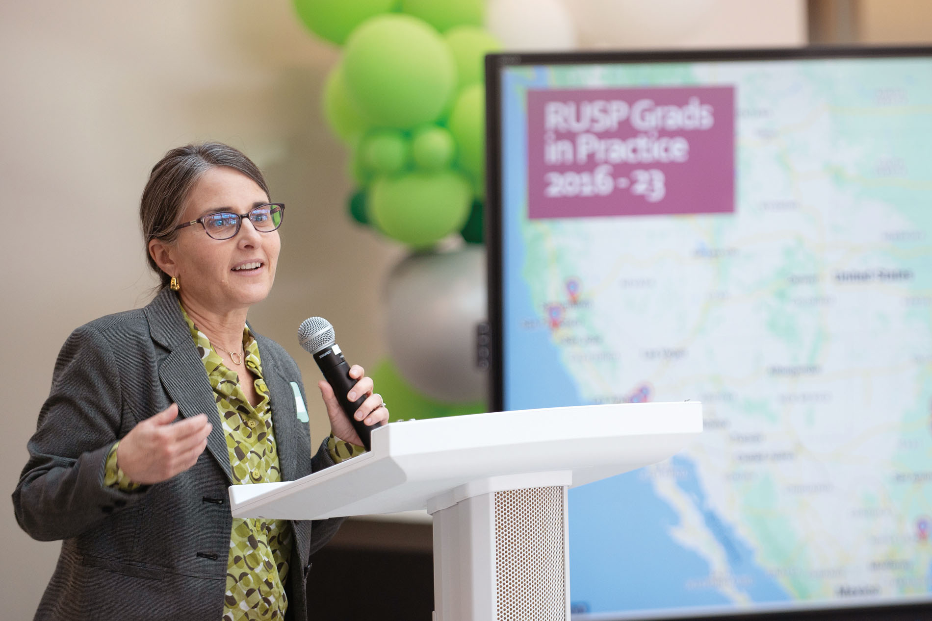 a woman with glasses and professional attire stands at a podium, speaking into a microphone