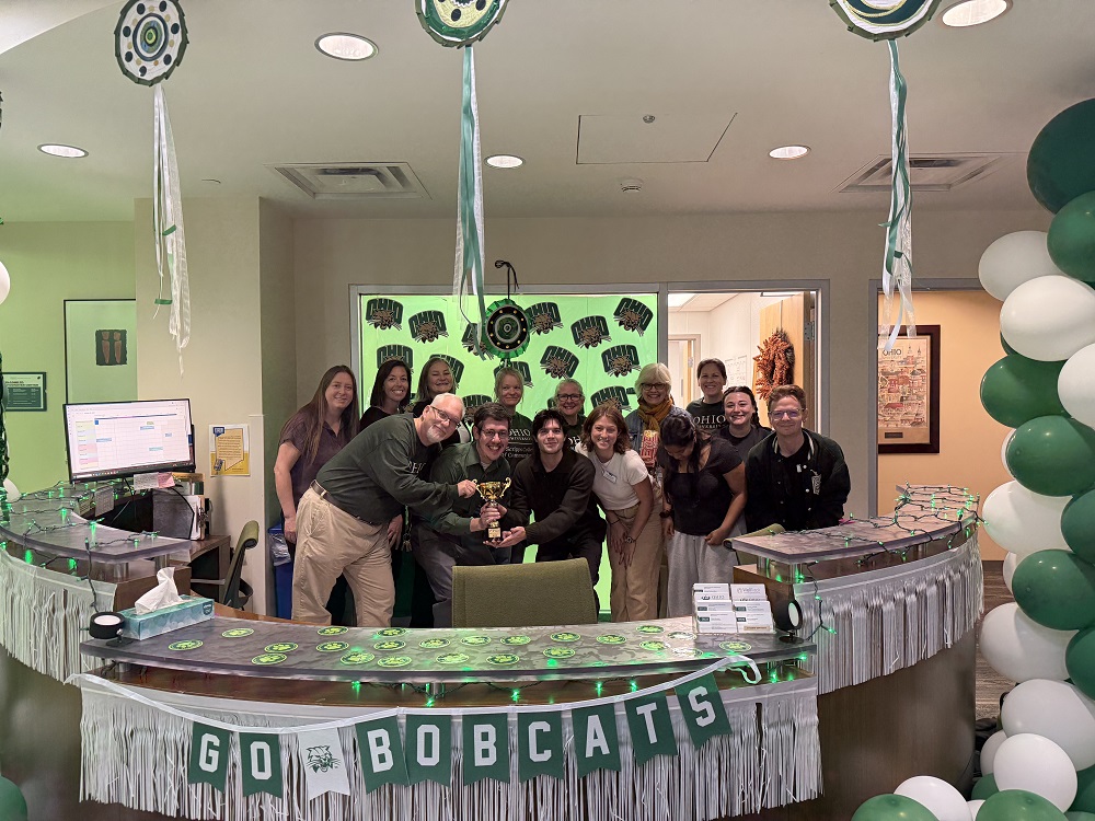 Scripps College faculty and staff hold their trophy while standing behind a desk in a room decorated for Homecoming