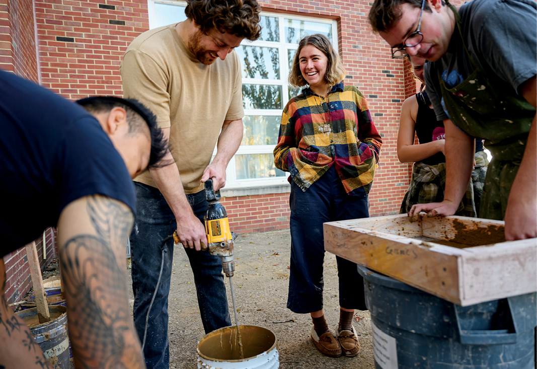 a professor in casual attire operates a drill with a mixer attachment to stir the contents of a 5-gallon bucket as several students look on