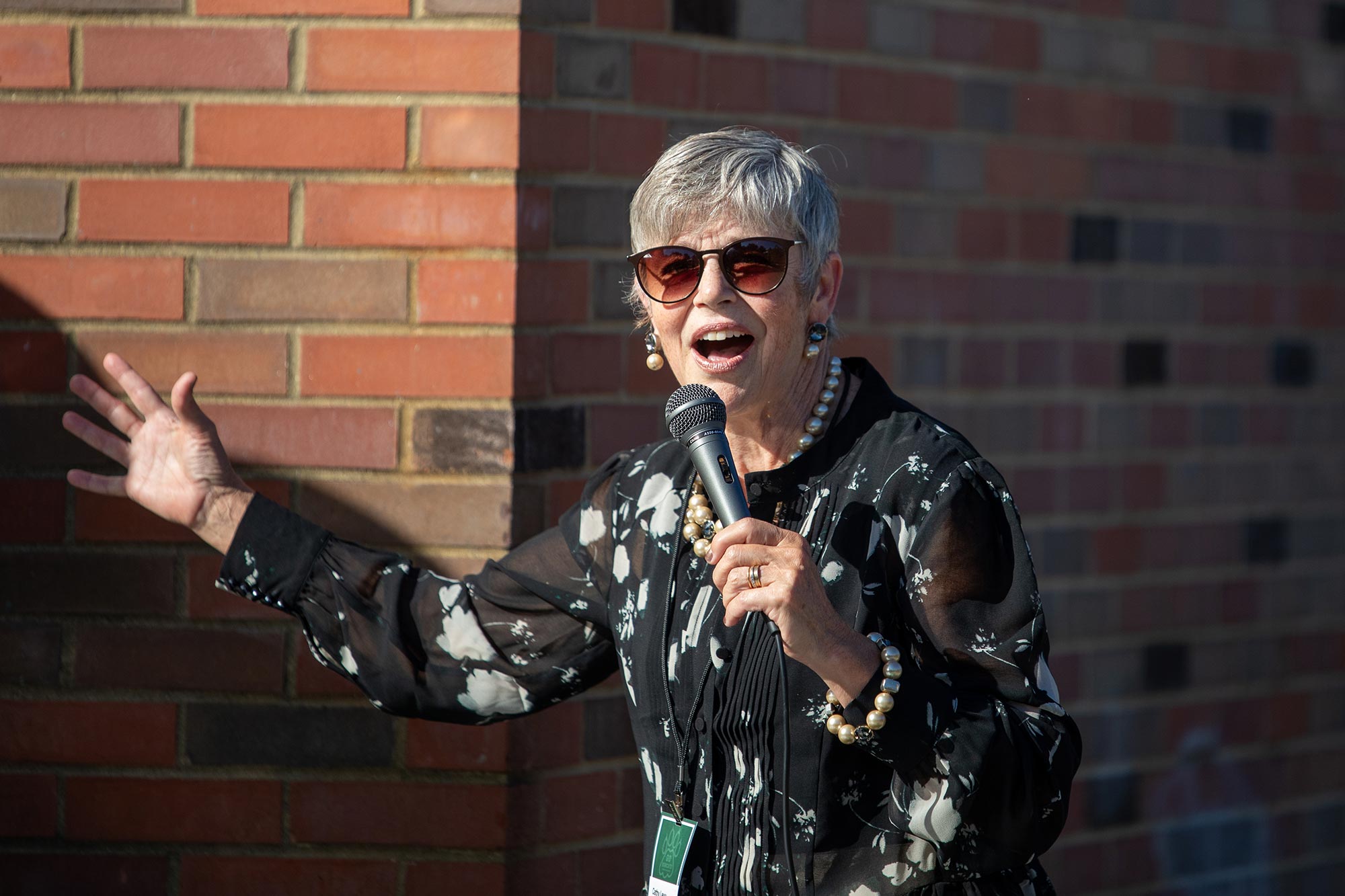 a late-middle-aged woman with gray hair, wearing sunglasses, a matching oversized pearl earring and necklace set, and a black shirt with floral print and sheer sleeves, gestures broadly while speaking into a microphone