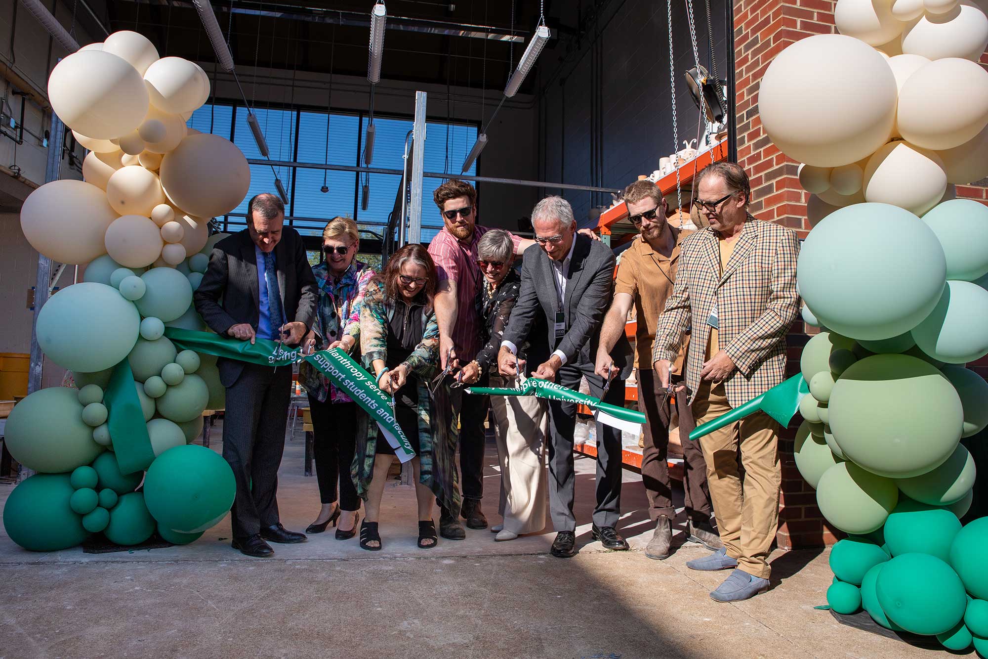 a group of adults in business casual attire and holding ceremonial silver scissors stand outdoors, cutting a green ribbon flanked by balloons