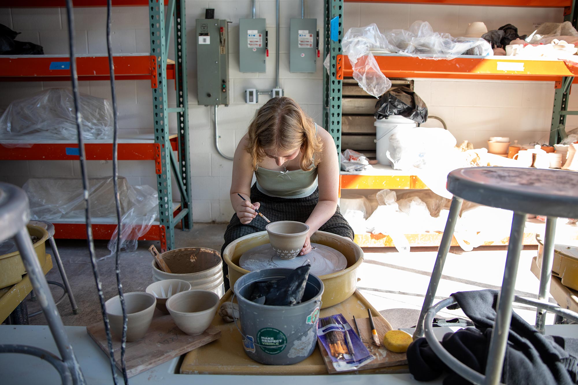 a female student sits in front of a throwing wheel, painting a clay pot with glaze