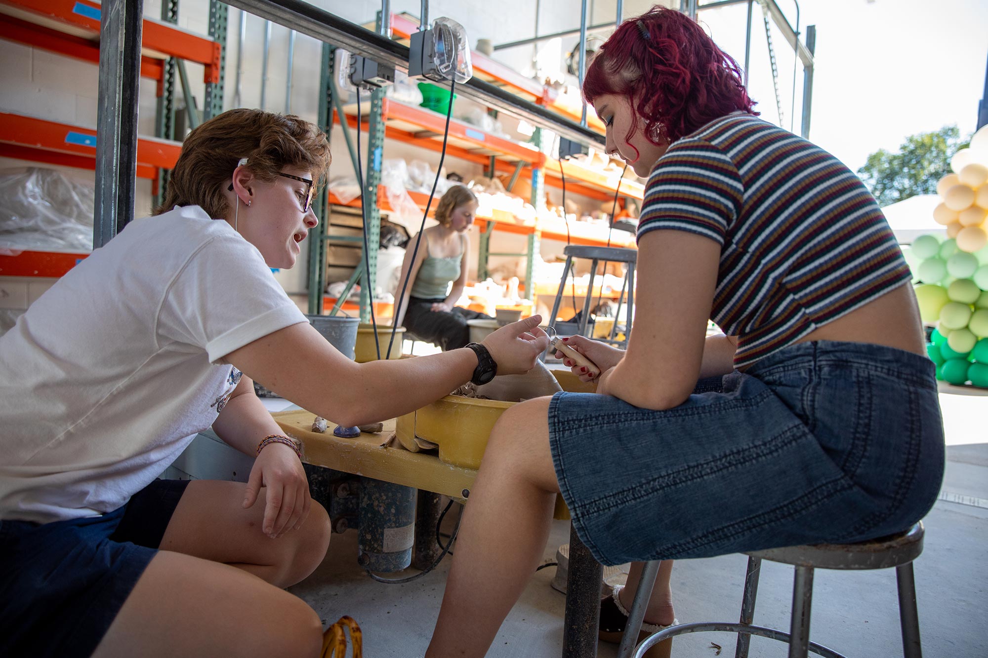 a student sits in front of a throwing wheel and a piece of clay while another student squats next to her, discussing technique 