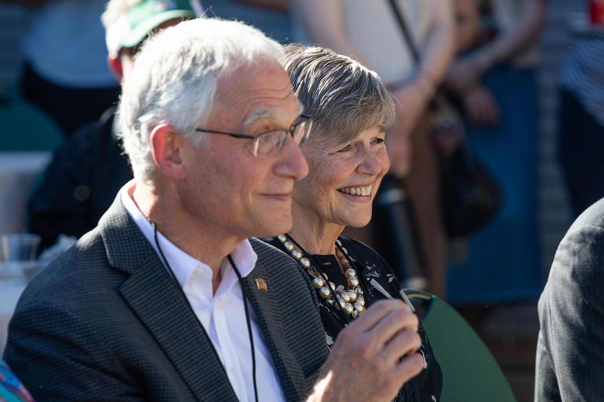 a late-middle-aged couple, both with short gray hair and wearing business casual attire; the man is wearing glasses and the woman has a large statement necklace, and both are smiling