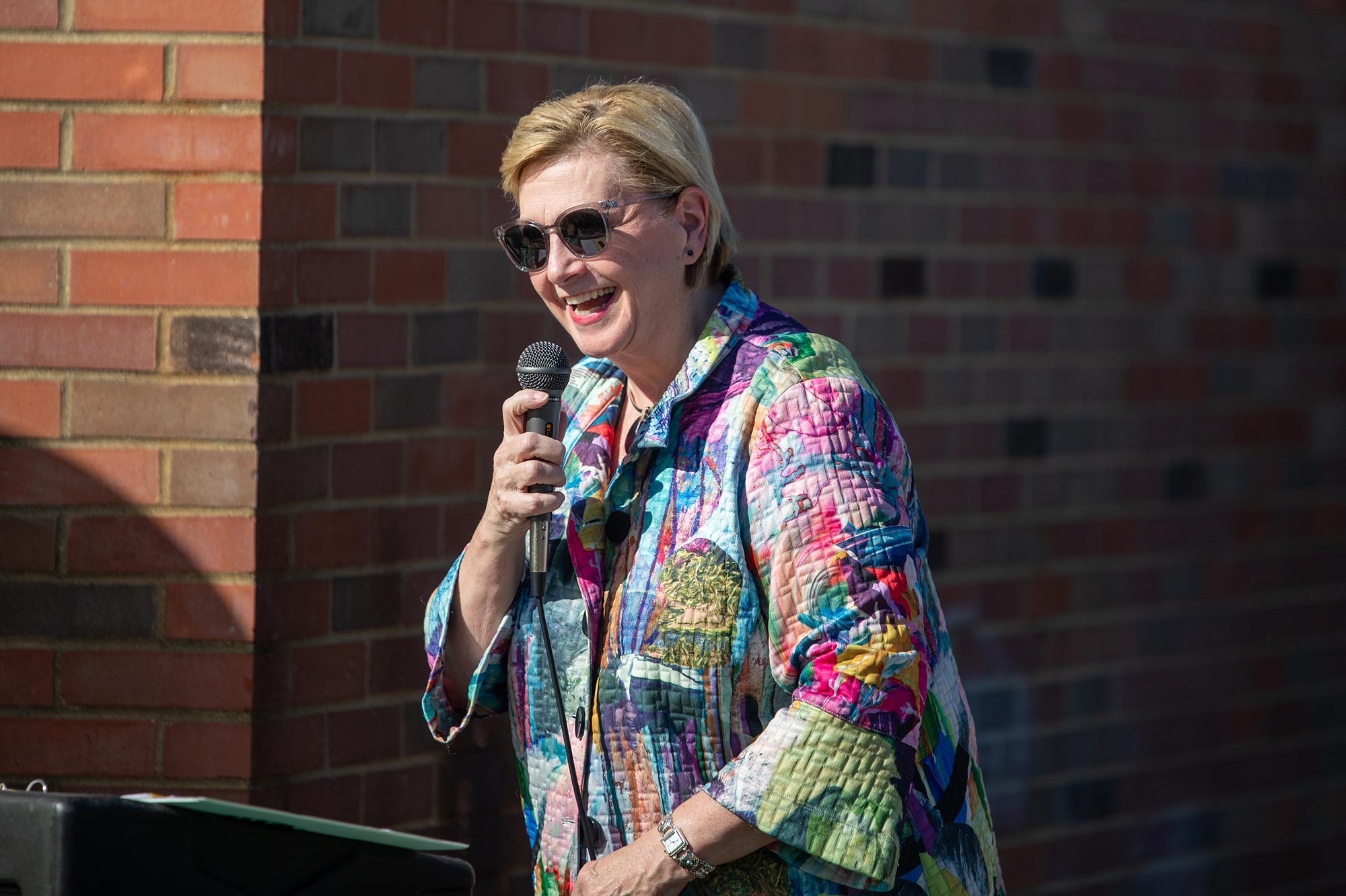 a blonde woman wearing sunglasses and a colorful shirt smiles while speaking into a microphone at an outdoor event