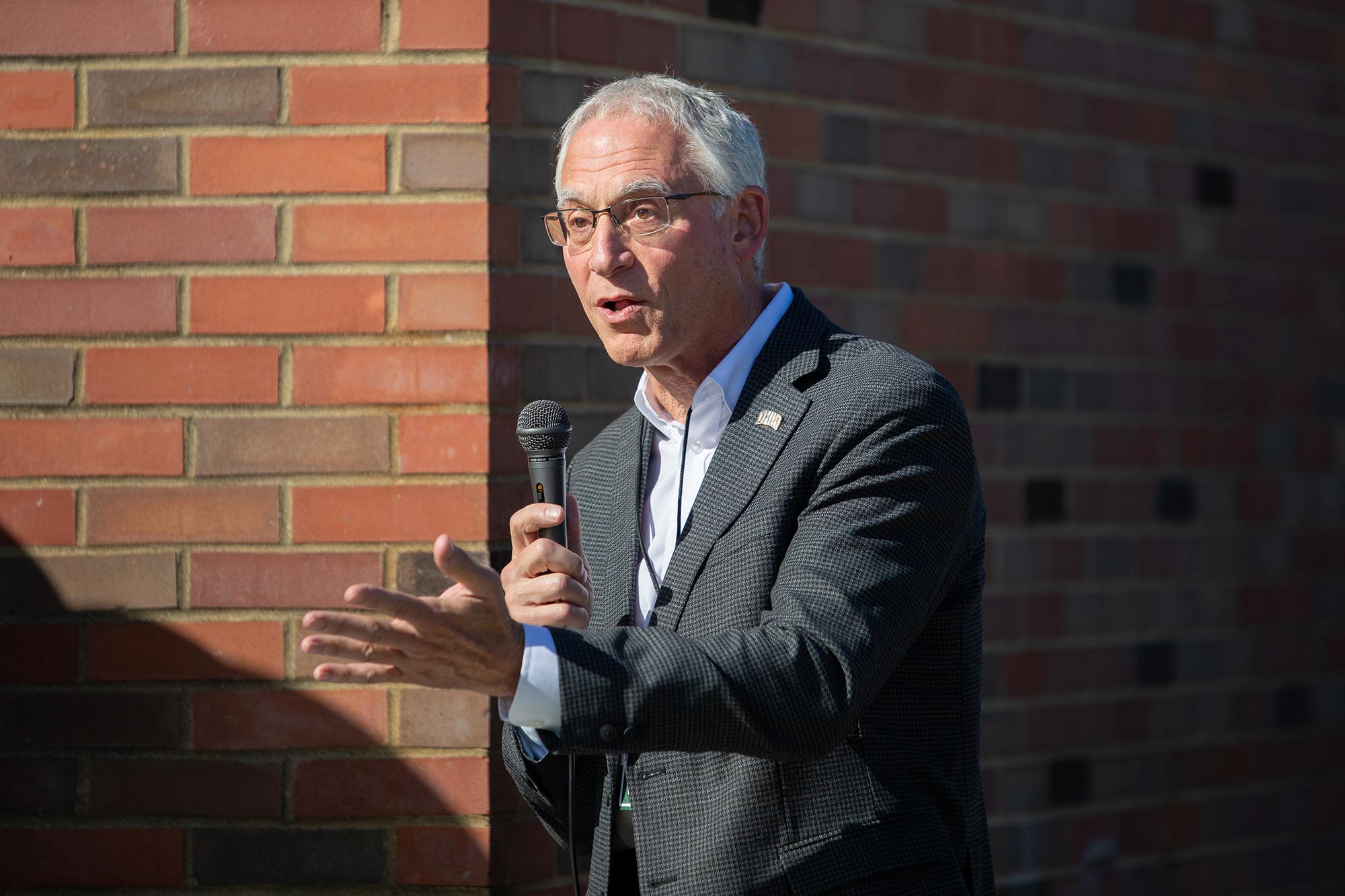 a late-middle-aged man with gray hair, wearing glasses, a blue button-down shirt and a dark herringbone pattern suit jacket, gestures and speaks into a microphone