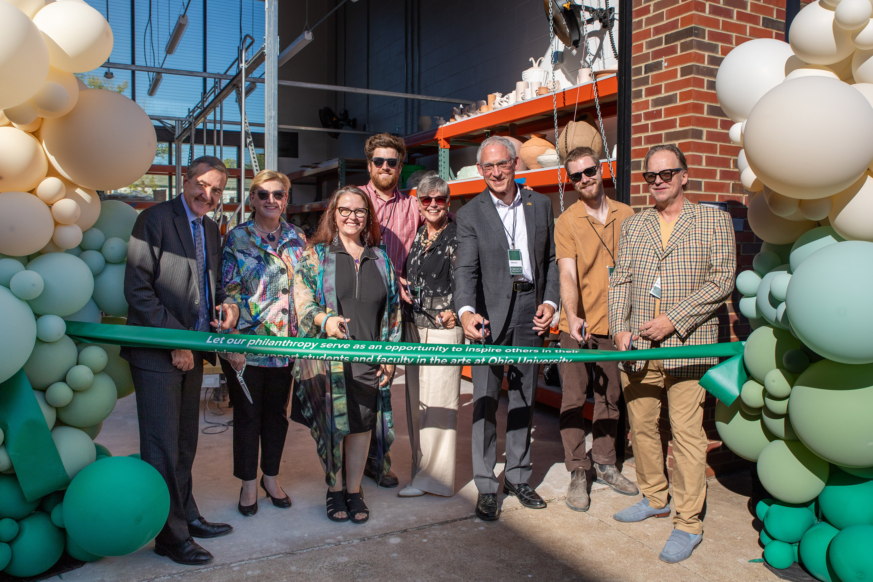 a group of adults in business casual attire and holding ceremonial silver scissors stand outdoors behind a green ribbon flanked by balloons