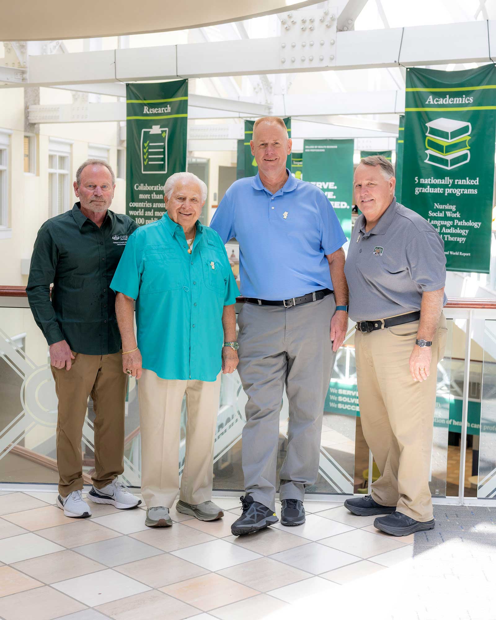 four men in business casual attire pose in front of green banners in a brightly lit atrium