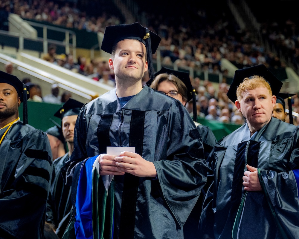 a graduate in cap and gown stands among other graduates, smiling