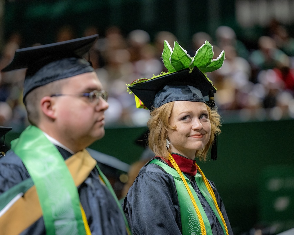 At the Commencement ceremony, a graduate whose cap is decorated with leaves smiles at someone beside her.