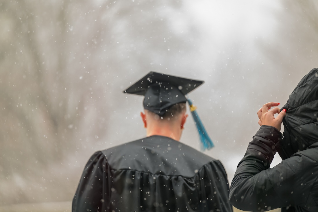 A graduate in cap and gown walks away from the camera, through snow