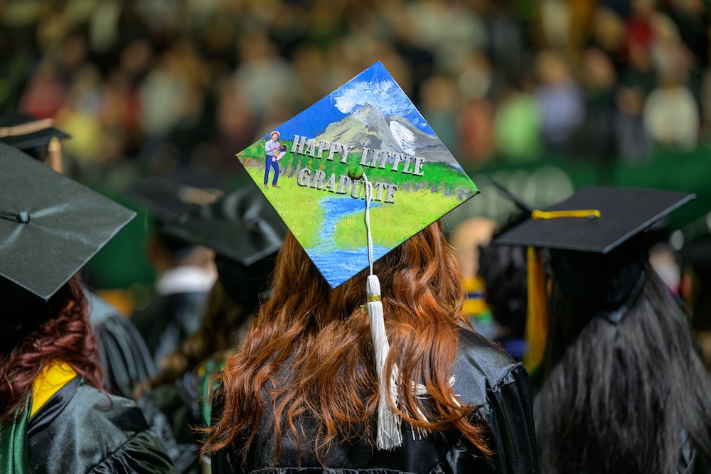 At the Commencement ceremony, a graduation cap features an image of a painter in a natures setting with the words "Happy Little Graduate"
