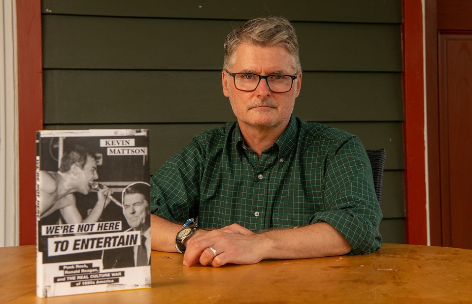 Kevin Mattson in a nice shirt sitting down at a wooden table next to his book.