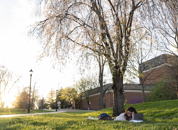 A student looks at a computer and papers while lying in the green grass under a colorful tree on a spring day at Ohio University