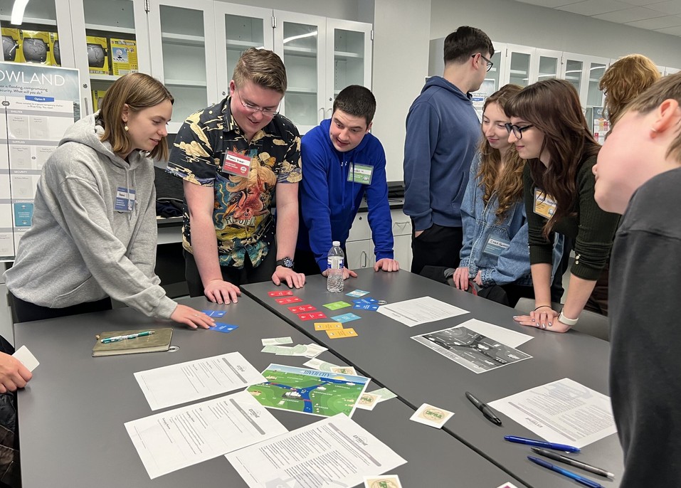Students examining paper sheets on a table in a classroom setting.