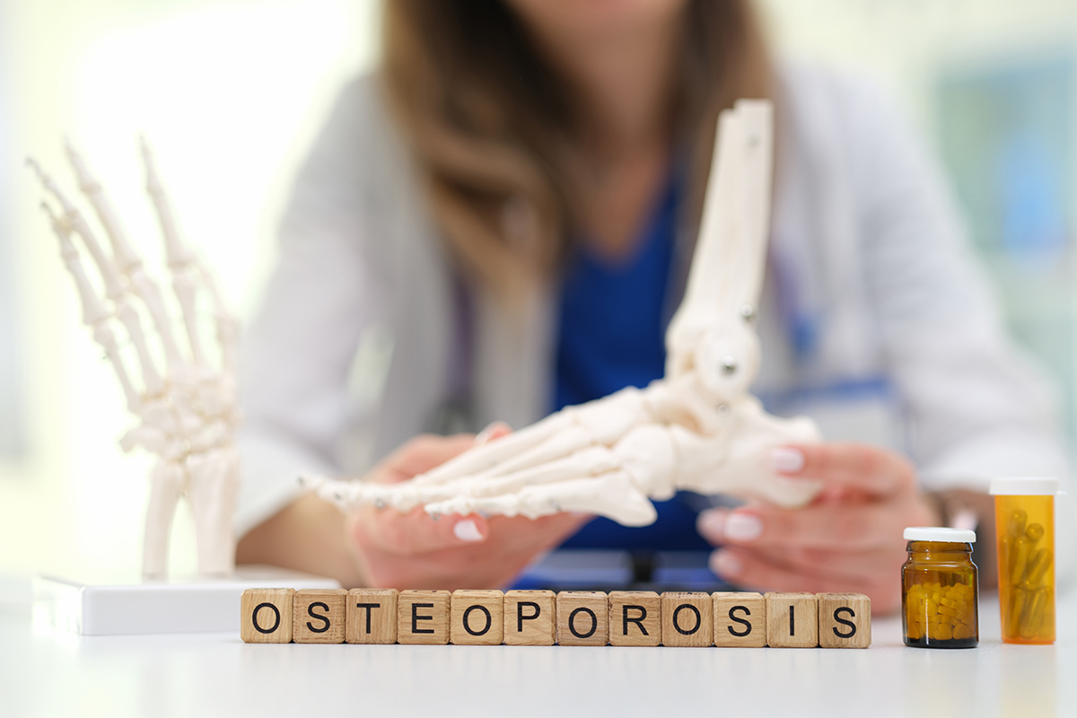 A physician examines skeletal models behind her desk. The letters OSTEOPOROSIS are spelled out in wooden blocks in front of her.
