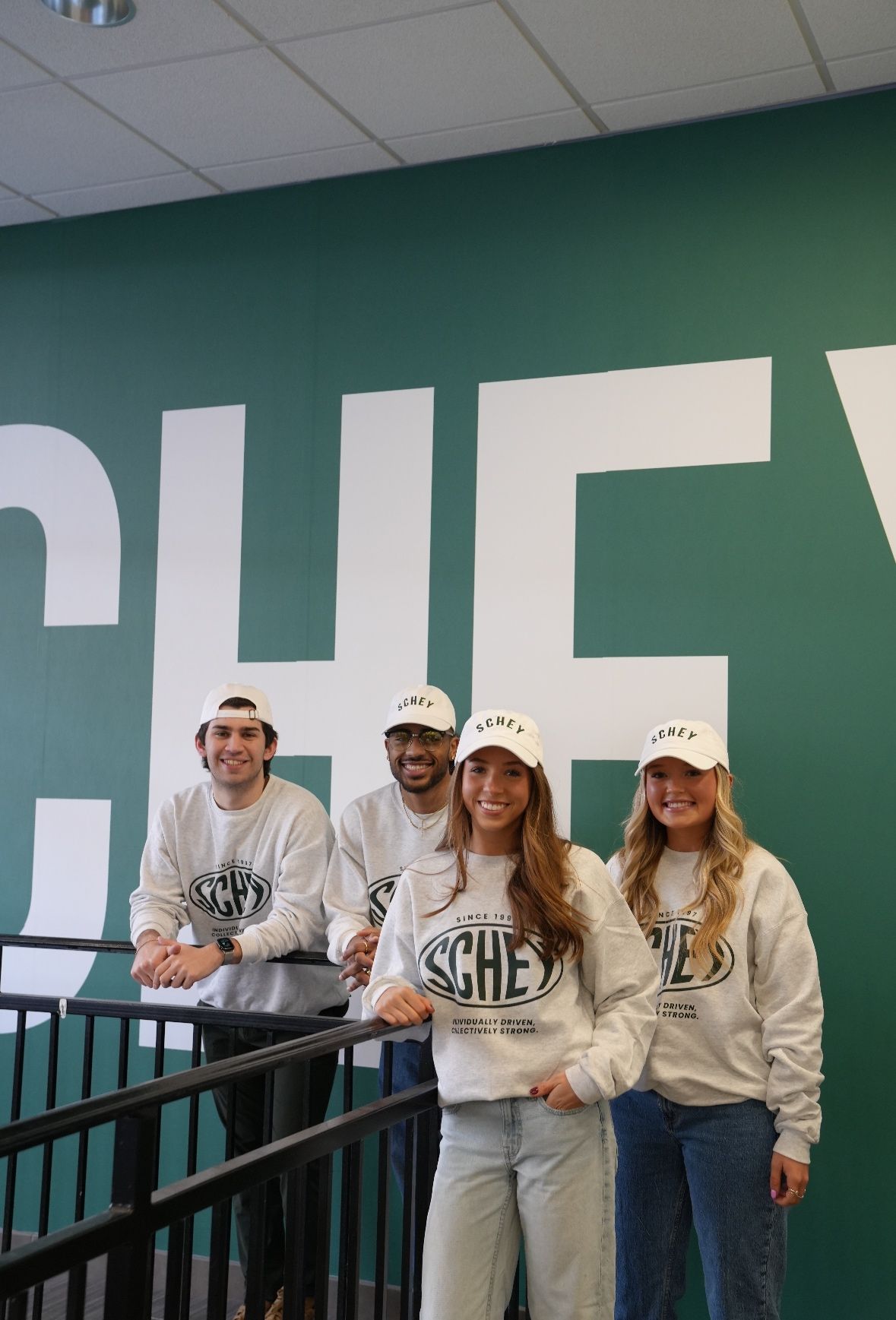 Four students in front of a giant wall that says "SCHEY" wearing Schey Sales Centre merch.